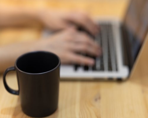 A close up of hands typing on a laptop over a desk with a warm cup of tea